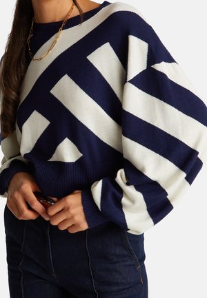 Femme portant un pull oversize rayé bleu marine et blanc avec de longues manches bouffantes, associé à un jean foncé et un collier chaîne en or.