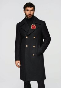 Schwarzer zweireihiger Wollmantel mit goldenen Knöpfen und einer dekorativen roten Herzbrosche, mit Reverskragen und Seitentaschen.