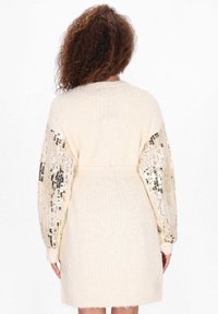 Kremowy sweter kardigan z długimi, połyskującymi rękawami obszytymi cekinami, z paskiem w talii i miękką teksturą. Wykończony żeberkowym splotem na korpusie.