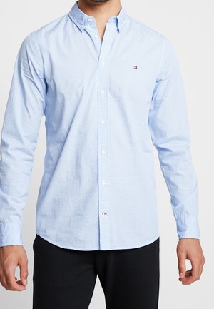 Mann trägt ein hellblaues, langärmliges Hemd mit feinem weißem Muster und kleinem Logo auf der Brust, kombiniert mit schwarzen Hosen.