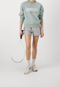 Sweatshirt verde chiaro con stampa "SPORTS CLUB", pantaloni corti grigi con strisce nere laterali, racchetta da tennis bianca e scarpe da ginnastica beige.