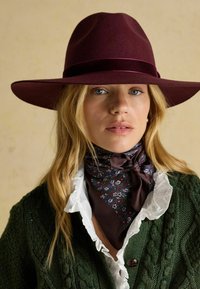 Pălărie cu boruri largi din fetru burgund cu o bandă asortată, purtată cu un fular maro cu model floral și un cardigan verde tricotat cu textură.