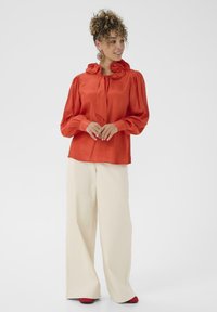 Blouse en satin rouge avec un col haut à volants et une nœud papillon. Manches longues avec des poignets froncés. Associée à un pantalon large crème.