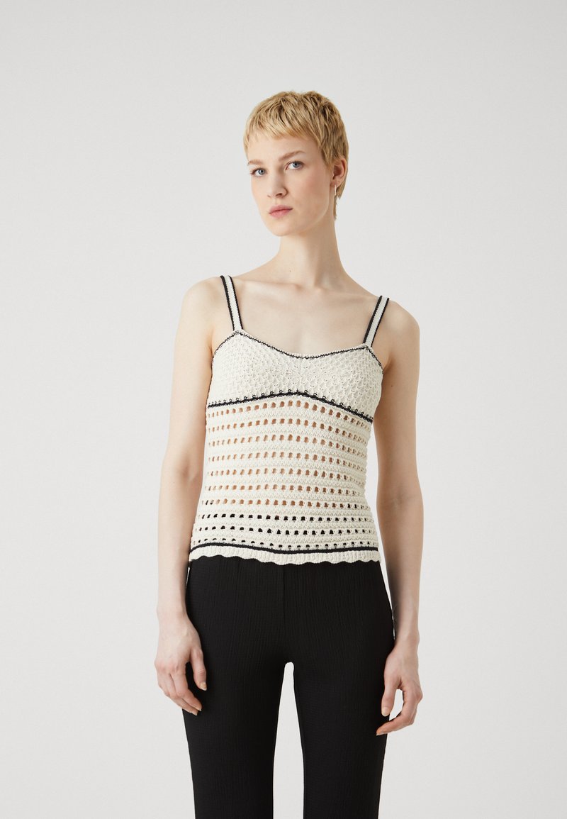 White crochet tank top with black trim and adjustable straps, featuring openwork patterns and a scalloped hem. Worn with black pants.