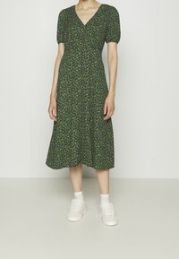 Robe midi verte à fleurs avec manches bouffantes, décolleté en V et corsage ajusté. Associée à des baskets blanches et des chaussettes de cheville.
