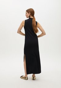 Mujer con cabello largo y rojo en un vestido maxi negro sin mangas con abertura lateral, pendientes de oro y zapatos planos metálicos dorados, de pie con las manos en las caderas.