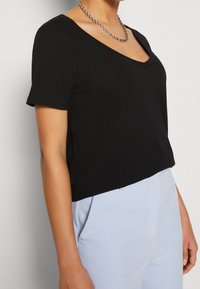 Kobieta w czarnej bluzce z dekoltem w serek i krótkim rękawem, z srebrnym naszyjnikiem i jasnoniebieskimi spodniami, na jednolitym jasnym tle.