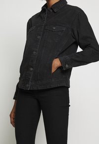 Persona con chaqueta de mezclilla negra con bolsillos en el pecho abotonados y pantalones negros, con una mano en el bolsillo lateral frente a un fondo liso.