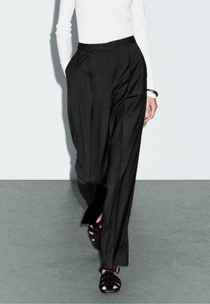 Schwarze karierte Hose mit taillierter Silhouette, ausgestattet mit Falten und Seitentaschen, kombiniert mit schwarzen Rückenriemen-Sandalen. Langes weißes Oberteil sichtbar.