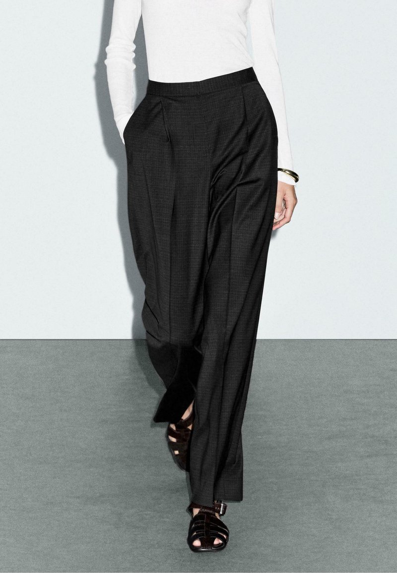 Black plaid trousers with a tailored silhouette, featuring pleats and side pockets, paired with black strappy sandals. White long-sleeve top visible.