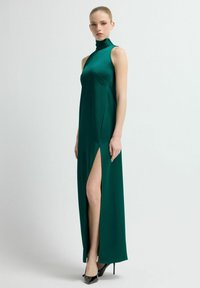 Dlouhé, přiléhavé zelené saténové šaty s vysokým límcem a bočním rozparkem na stehnu. Hladká textura a elegantní design spojené s černými lodičkami na vysokém podpatku.