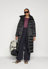 Casaco acolchoado preto com capuz, silhueta longa, textura acolchoada. Combinado com uma camisola bordô, calças pretas e uma mochila cinza. Calçado castanho.