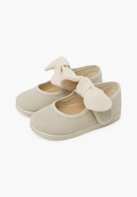 Zapatos de tela beige con un diseño texturizado, punta redondeada y un gran detalle de lazo atado. Cuentan con una correa ajustable segura.
