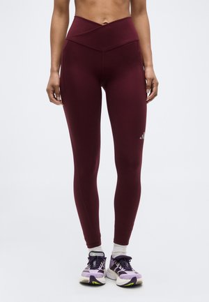 Donna che indossa leggings bordeaux a vita alta e scarpe da ginnastica viola con calzini bianchi, in piedi su uno sfondo chiaro e semplice.