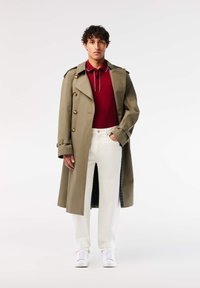 Kaki trenchcoat med guldknappar, tillsammans med en röd polo och vita byxor. Vita sneakers fulländar outfiten.