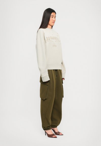 Woman with long dark hair wearing beige sweatshirt, olive green cargo pants with large pockets, and brown open-toe high heels, standing sideways.