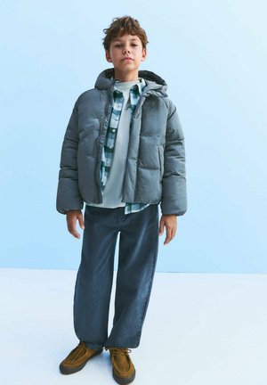 Garçon portant une doudoune grise par-dessus une chemise à carreaux verte et un t-shirt blanc, pantalon large foncé et chaussures marron, debout devant un fond bleu clair.