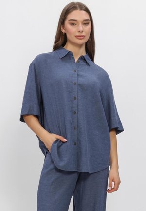 Femme portant une chemise bleue ample à manches courtes boutonnée et un pantalon assorti, debout avec une main dans la poche devant un fond uni.