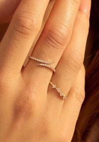 Deux bagues en or sur une main : l'une présente un motif ondulé avec de petits diamants ; l'autre a trois diamants carrés disposés en ligne.