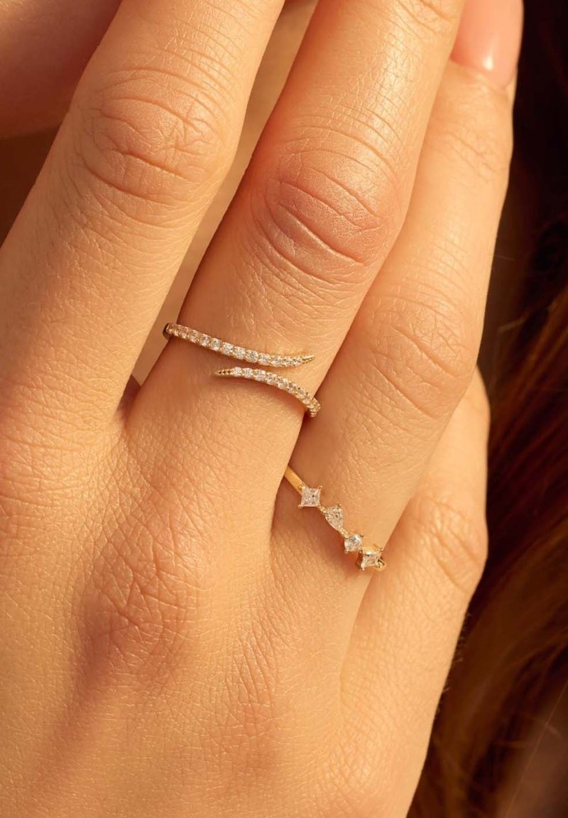 Deux bagues en or sur une main : l'une présente un motif ondulé avec de petits diamants ; l'autre a trois diamants carrés disposés en ligne.