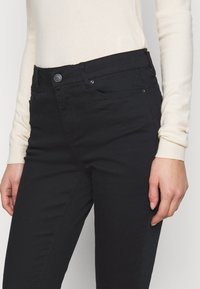 Los jeans negros presentan un diseño ajustado con cierre de botón y cremallera, presillas para cinturón y detalles de costura sutil. El material parece elástico y suave.