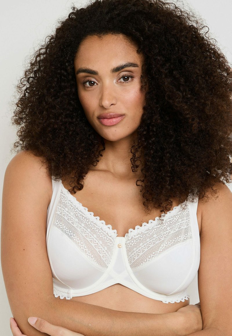 Brasile bianca con dettagli in pizzo, bordi ondulati e un bottone al centro. Presenta spalline regolabili e una texture liscia e morbida.