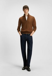 Maglione marrone a zip realizzato in tessuto testurizzato, abbinato a pantaloni in denim scuri. L'outfit è completato da mocassini neri.