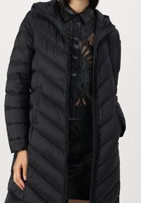 Abrigo acolchado negro con un patrón de chevron, cierre de cremallera y capucha. El forro es suave, complementando un detalle floral negro translúcido debajo.