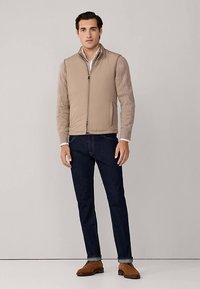 Béžová prošívaná vesta s žebrovanými dlouhými rukávy, předním zipem a bočními kapsami, doplněná tmavými džínami a hnědými botami. Jednoduchý design.