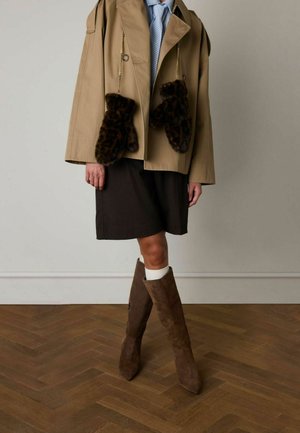 Tan jas met oversized pasvorm, voorzien van faux fur wanten met luipaardprint. Gecombineerd met een bruine rok en bruine suède kniehoge laarzen.