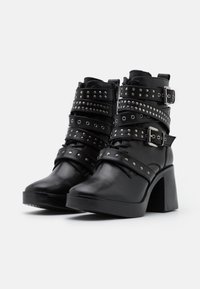 Botas negras de tobillo con un tacón grueso, que presentan múltiples correas con tachuelas y hebillas de plata, hechas de cuero liso con un acabado mate.