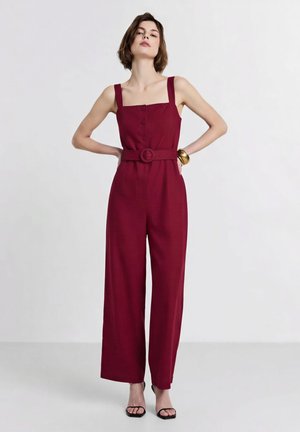 Frau mit kurzen braunen Haaren, die einen ärmellosen bordeauxfarbenen Jumpsuit mit weitem Bein und passendem Gürtel trägt, steht mit den Händen in der Hüfte.
