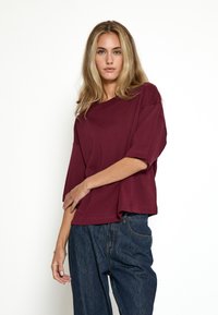 Camiseta de gran tamaño en burdeos, fabricada en algodón suave, con cuello redondo y mangas cortas, combinada con pantalones de mezclilla oscuros.