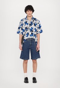 Cămașă cu mânecă scurtă, albă, cu imprimeu de animale albastre, asociată cu șorturi din denim albastru, de lungime până la genunchi, și pantofi negri slip-on, completată cu o geantă crossbody.