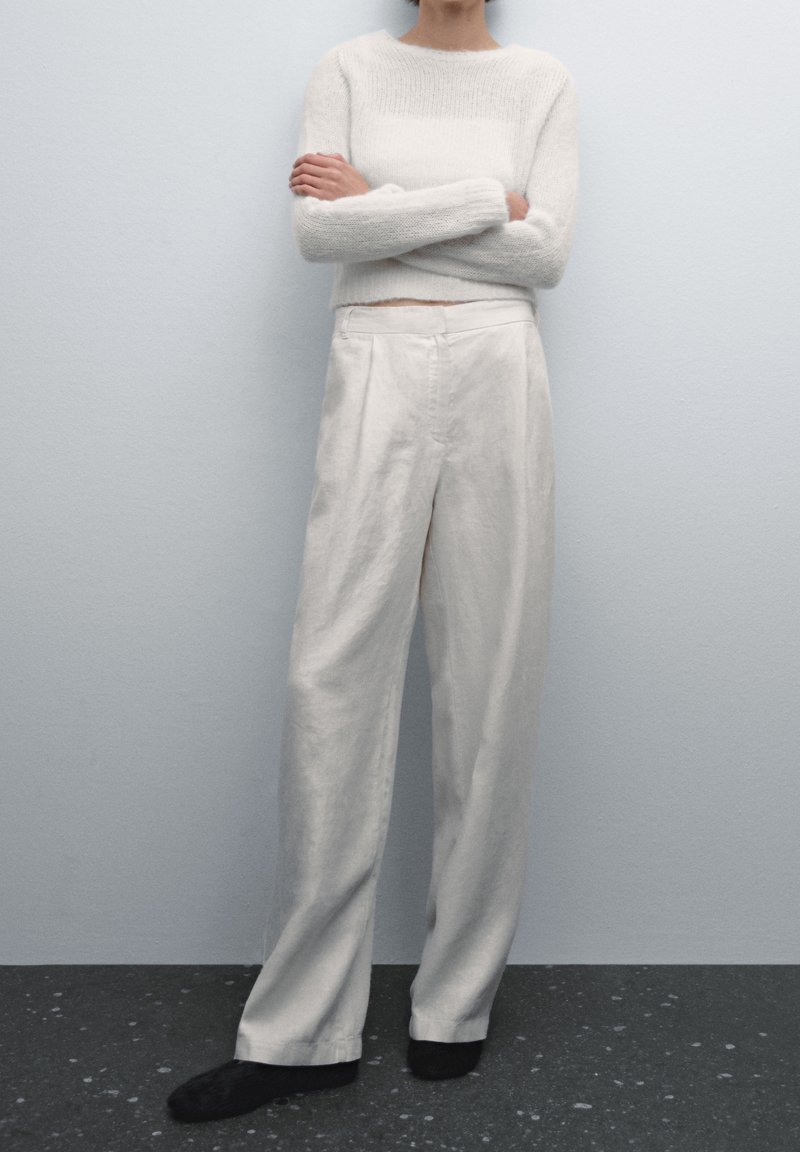 Personne debout les bras croisés, portant un haut blanc duveteux à manches longues, un pantalon blanc ample et des chaussures noires sur un fond clair uni.