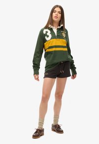 Maillot de rugby vert à manches longues avec une bande jaune et le numéro 3, associé à un short noir. Stylisé avec des chaussettes beiges et des chaussures marron.