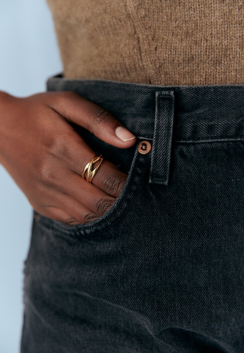 Svarta jeans med en strukturerad yta och en synlig kopparknapp, med en hand som bär en glänsande, sammanflätad guldring.