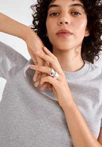 Young person with curly hair wearing a light gray T-shirt, showing two silver rings on crossed fingers near their face.