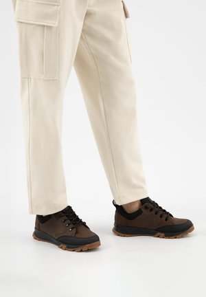 Personne portant un pantalon cargo beige et des chaussures de randonnée marron avec semelles et lacets noirs, se tenant sur une surface blanche.
