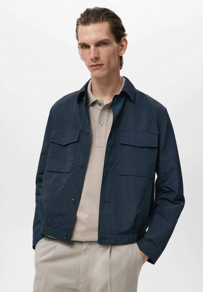 Chaqueta azul marino de manga larga, con cierre de botón y dos bolsillos frontales, llevada sobre un polo beige. Textura suave, diseño informal.