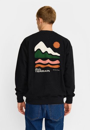 Persona que lleva una sudadera negra con gráficos de montaña, sol y ola y texto "ALL TERRAIN" en la espalda, combinada con vaqueros azules.