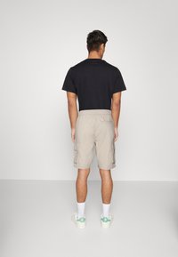Hombre de pie frente a una pared, vistiendo una camiseta negra, pantalones cortos beige de cargo, calcetines blancos y zapatillas blancas con detalles verdes.