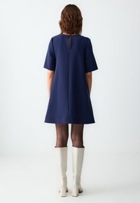 Vestito a tunica blu navy a maniche corte con dettaglio a chiave sulla schiena, abbinato a stivali bianchi sopra il ginocchio e collant neri. Tessuto morbido, vestibilità ampia.
