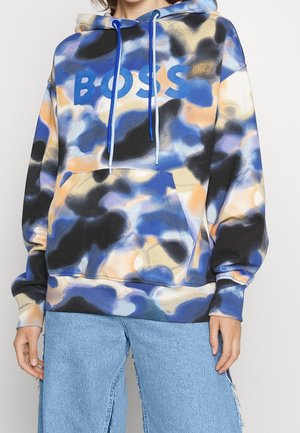 Personne portant un sweat à capuche camouflage bleu, noir et beige avec le texte "BOSS", associé à un jean large bleu clair.