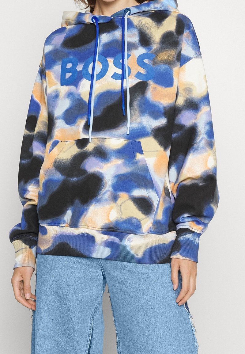 Personne portant un sweat à capuche camouflage bleu, noir et beige avec le texte "BOSS", associé à un jean large bleu clair.