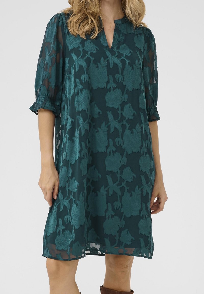 Robe ample vert foncé longueur genou avec broderie florale, manches transparentes longueur coude et légers volants aux poignets sur une femme debout.