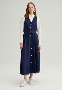 Robe bleu marine avec un haut de vêtement ajusté, des boutons décoratifs et une jupe évasée. Portée avec des baskets blanches et des manches longues sous le haut.