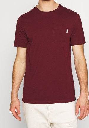 Mand iført en ensfarvet bordeaux kortærmet t-shirt med en lille brystlomme og hvide bukser, stående mod en neutral baggrund.