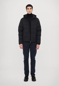 Black puffer jacket with quilted pattern, zip closure, and attached hood, worn over a black turtleneck. Paired with navy trousers and black shoes.