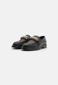 Black leather loafers featuring a prominent gold chain accent on the front, with a smooth texture and rounded toe shape, set on a low heel.